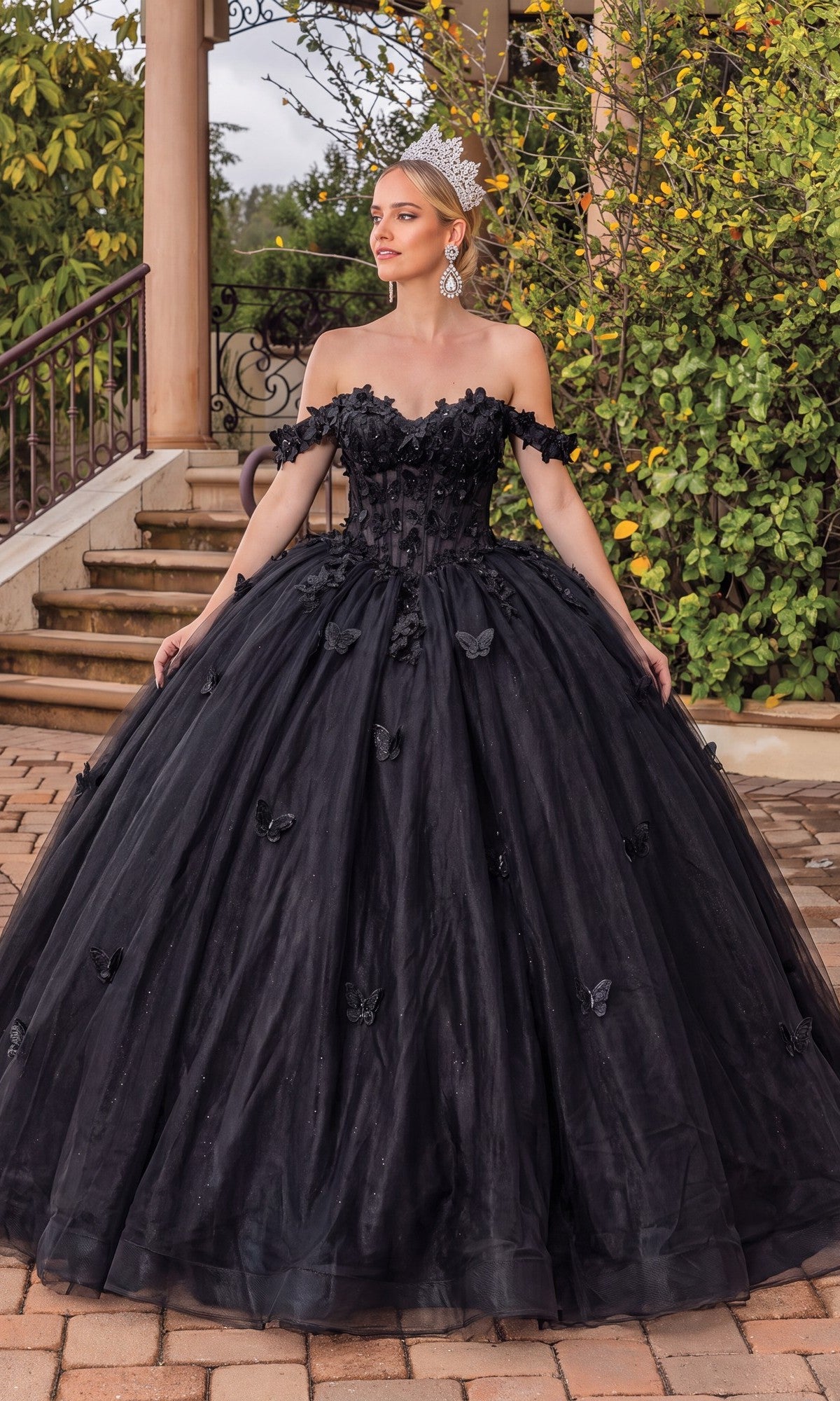 A woman wearing the Quinceanera Dress 1826 by Dancing Queen in black, featuring elegant lace applique and an off-the-shoulder ball gown silhouette, stands outdoors on a stone patio near steps, surrounded by greenery and adorned with a tiara.